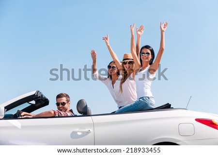 Spending great time together. Group of young happy people enjoying road trip in their white convertible while girls raising arms and smiling