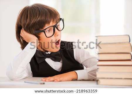 His favorite books. Excited young boy in shirt and bow tie sitting at the table and looking at book stack