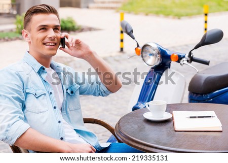 Good talk with friend. Relaxed young man talking on the mobile phone and smiling while sitting in sidewalk cafe