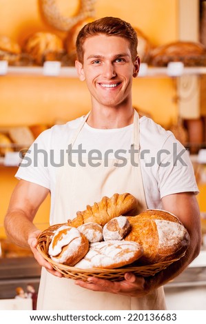 Proud of his baked goods. Handsome young man in apron holding basket with baked goods and smiling while standing in bakery shop
