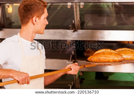 Fresh baked bread. Confident young man in apron taking the fresh baked bread from oven