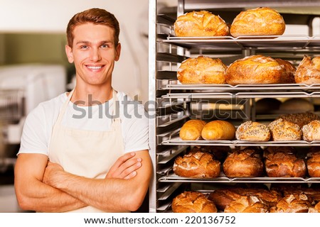 I love my job! Handsome young baker in apron keeping arms crossed and smiling while leaning at the tray with fresh baked bread