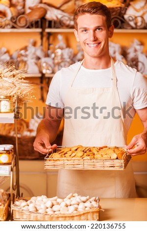 The freshest cookies for you. Handsome young man in apron holding tray with cookies and smiling while standing in bakery shop