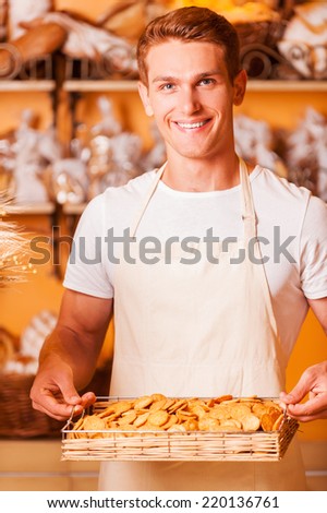 The fresh baked cookies for you. Handsome young man in apron holding tray with cookies and smiling while standing in bakery shop