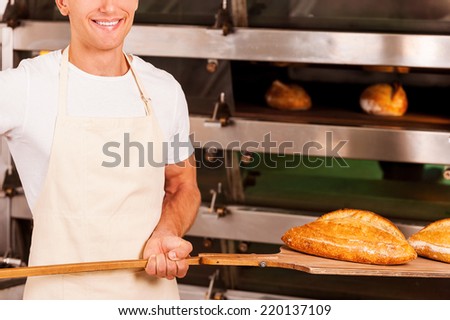 Fresh baked bread for you. Cropped image of confident young man in apron taking fresh baked bread from oven and smiling