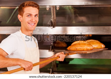 Confident baker. Confident young man in apron taking fresh baked bread from oven and smiling