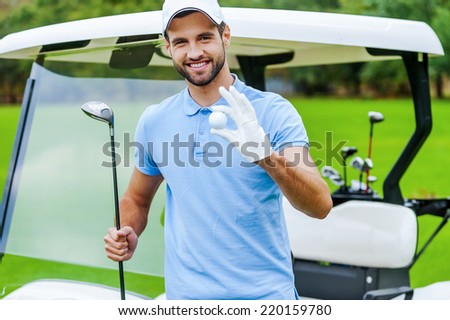 Only the best golf equipment! Handsome young smiling man holding golf ball and driver while standing near the golf cart and on golf course