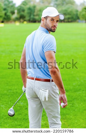 Confident golfer. Rear view of confident golfer holding golf ball and driver while standing on golf course and looking over shoulder
