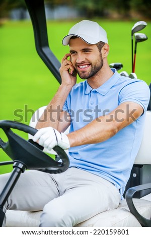 I will meet you on green! Handsome young smiling man driving a golf cart and talking on the mobile phone