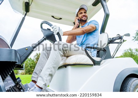 I am on my way to next hole! Handsome young smiling man driving a golf cart and talking on the mobile phone
