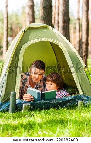 Spending good time outdoors. Father and son reading book while lying in tent together