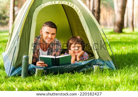 Camping together is fun. Father and son reading book and smiling while lying in tent together