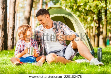 Outdoor adventure. Cheerful father and son sitting near the tent and drinking hot tea from metal cups while camping in the forest together