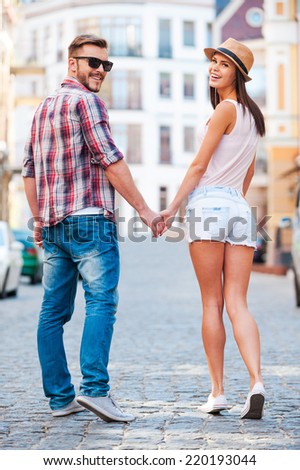 Enjoying their time together. Rear view of beautiful young loving couple walking by the street and looking over shoulder