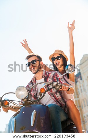 Riding with fun. Beautiful young couple riding scooter together while happy woman raising arms and smiling