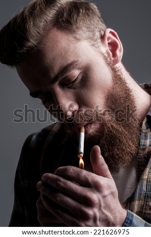 Lighting up cigarette. Handsome young bearded man lighting up a cigarette while standing against grey background