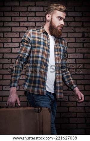 Ready to go. Handsome young bearded man carrying suitcase while walking away with brick wall as background