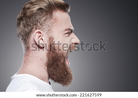 Unleashing his emotions. Side view of furious young bearded man shouting while standing against grey background