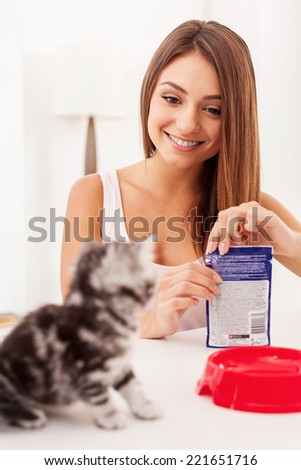 Are you hungry? Beautiful young woman opening a pack with cat food and smiling while little kitten waiting while sitting on foreground