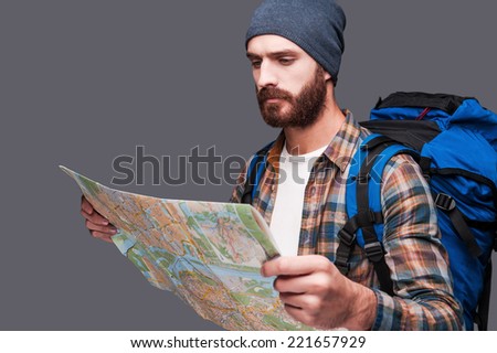 Tourist examining map. Handsome young bearded man carrying backpack and examining map while standing against grey background
