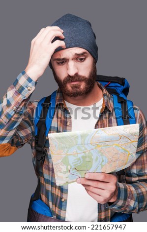 Lost in somewhere. Frustrated young bearded man carrying backpack and examining map while scratching head and standing against grey background