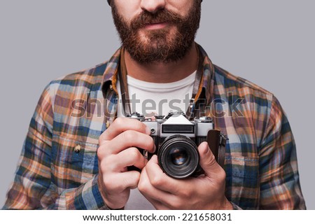 Smile to the camera! Close-up of young bearded man holding old-fashioned camera while standing against grey background