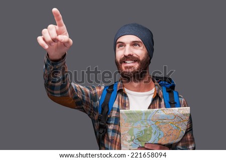 I know the right direction! Happy young bearded man with backpack holding map and pointing away while standing against grey background