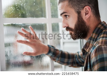 Rainy mood. Thoughtful young bearded man touching the window and looking through it