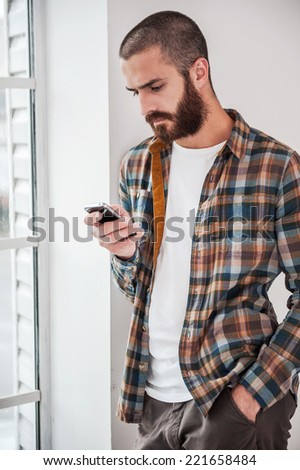 Typing text message. Confident young bearded man typing message on his smart phone while standing near the window