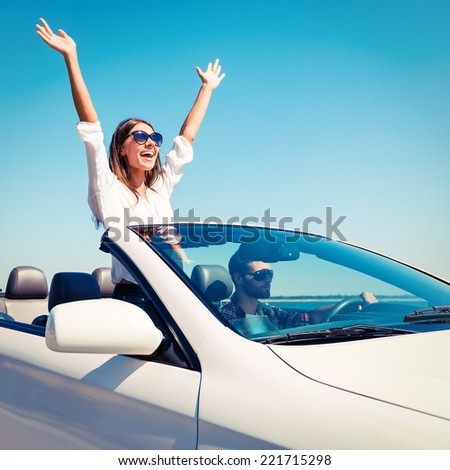 Couple in convertible. Happy young couple enjoying road trip in their convertible while woman raising arms and smiling