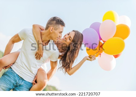 Bright love. Low angle view of young man carrying beautiful cheerful girl with colorful balloons on his shoulders