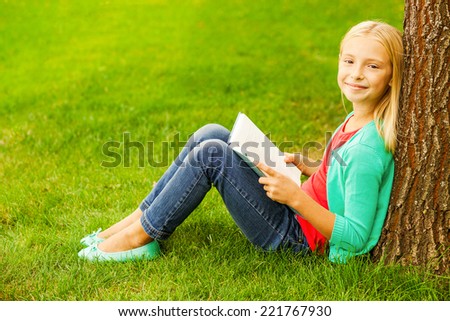 Little bookworm. Cute little blond hair girl holding book and smiling while sitting on green grass and leaning at the tree