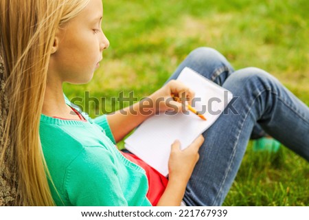 Performing her creativity. Cute little blond hair girl drawing something at her note pad while sitting on green grass and leaning at the tree