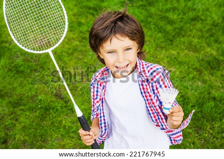 Little champion. Top view of happy little boy holding badminton racket and shuttlecock while standing on green grass