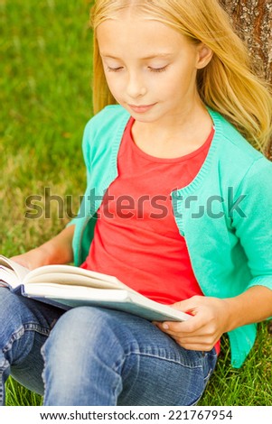 Reading her favorite book. Top view of cute little blond hair girl reading book while sitting on green grass and leaning at the tree