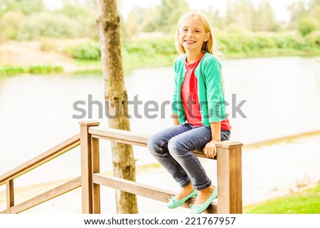 So good to do nothing. Cute little girl sitting at the wooden railing and smiling with river in the background