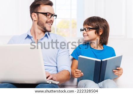 As father as son. Happy father working on laptop and looking at his little son sitting close to him and reading a book