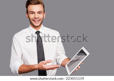 Keep up with technological progress. Cheerful young man holding a digital tablet and pointing it while standing against grey background