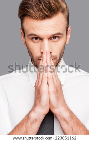 It is urgent question. Thoughtful young man in formalwear holding hands clasped near face while standing against grey background