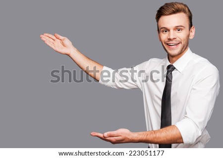 Positive selling. Confident young man in shirt looking at camera and pointing away while standing against grey background