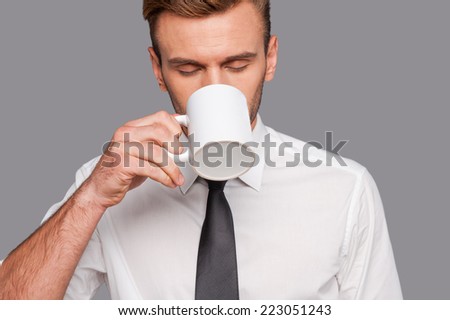 Finally coffee break . Tired young man in formalwear holding coffee cup and drinking it while standing against grey background