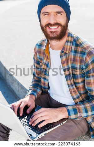 Such a good day for working outdoors!  Handsome young bearded man smiling while working on laptop and looking at camera