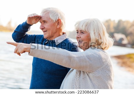 Look over there! Happy senior couple standing on the quayside together while woman pointing away and smiling