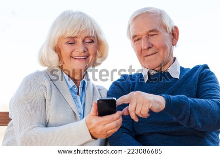 Technologies become easier. Cheerful senior couple looking at the mobile phone and smiling while sitting on the park bench together