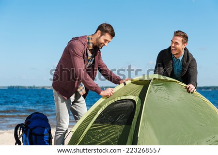 Setting up a tent. Two cheerful young men setting up a tent while standing at the riverbank