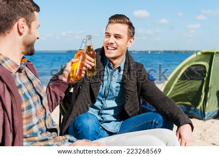 Cheers! Two happy men cheering with beer and smiling while sitting near the tent together
