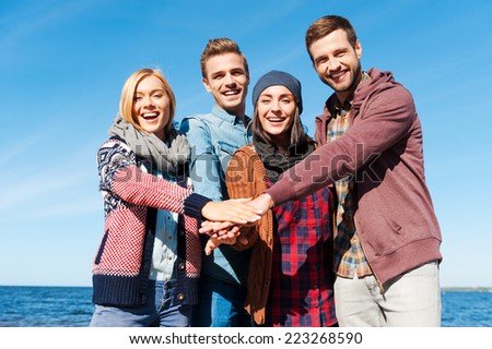 We are the best friends. Four young happy people bonding to each other and holding hands together while standing on the beach with river in the background