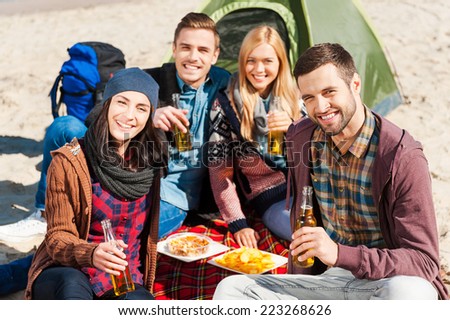 Spending time with friends. Group of young cheerful people drinking beer and talking while camping at the riverbank