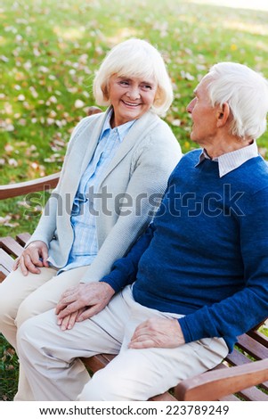 Relaxing in park together. Top view of happy senior couple holding hands and looking at each other while sitting on the bench in park together