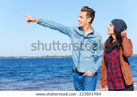 Look over there! Beautiful young couple walking by the beach together while handsome man pointing away and smiling